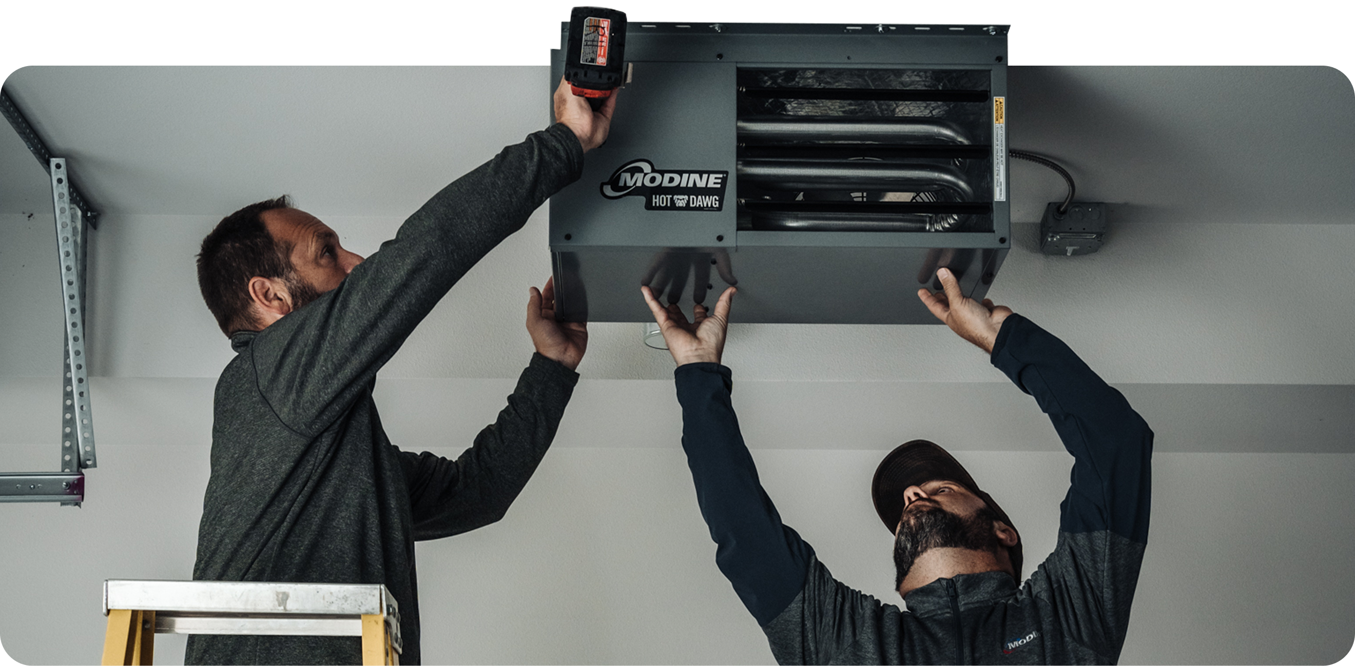 Two contractors hanging a Modine Hot Dawg unit heater from a garage ceiling